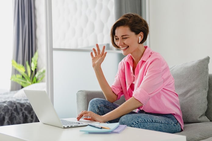 woman in pink shirt sitting relaxed on sofa at home at table working online on laptop