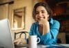 Cheerful attractive young woman enjoying distant work, sitting at desk using portable computer, drinking coffee. Pretty female blogger working from home, uploading video on her channel, smiling