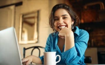 Cheerful attractive young woman enjoying distant work, sitting at desk using portable computer, drinking coffee. Pretty female blogger working from home, uploading video on her channel, smiling