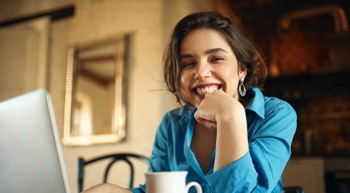 Cheerful attractive young woman enjoying distant work, sitting at desk using portable computer, drinking coffee. Pretty female blogger working from home, uploading video on her channel, smiling