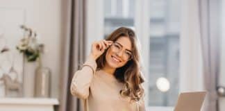 Cute girl in beige shirt touching glasses and holding laptop with smile. Indoor photo of beautiful female student preparing for lesson with computer at home..