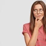 Photo of unsure doubtful young woman holds chin, looks right doubtfully, reconsiders something, feels hesitant, dressed in casual striped t shirt, poses against white background with copy space