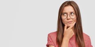 Photo of unsure doubtful young woman holds chin, looks right doubtfully, reconsiders something, feels hesitant, dressed in casual striped t shirt, poses against white background with copy space
