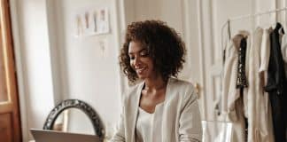 Attractive curly dark skinned woman works in laptop and sits by desk with coffee cup on it. Happy d