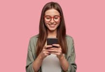 Satisfied lovely woman holds modern cell phone, texts with friend online in chat, dressed in stylish clothes, poses against pink wall, connected to high speed internet. Shot of teenager with device