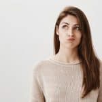 Student is bored making up ideas what to do. Portrait of carefree dreamy caucasian girl looking up with puckered lips, thinking how to entertain herself, wanting to call friend over gray background