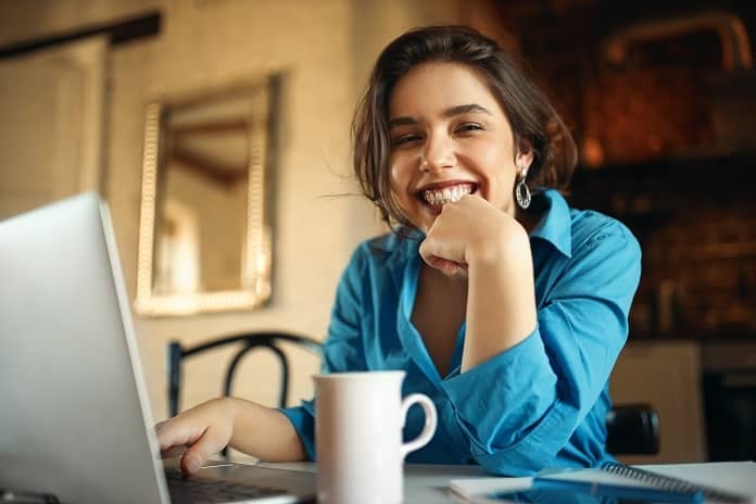 Cheerful attractive young woman enjoying distant work, sitting at desk using portable computer, drinking coffee. Pretty female blogger working from home, uploading video on her channel, smiling