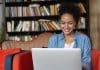 People, technology and education concept. Happy pretty dark skinned female in jean shirt sitting at sofa using laptop, typing something and browsing internet while being in reading hall or library.