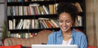 People, technology and education concept. Happy pretty dark skinned female in jean shirt sitting at sofa using laptop, typing something and browsing internet while being in reading hall or library.