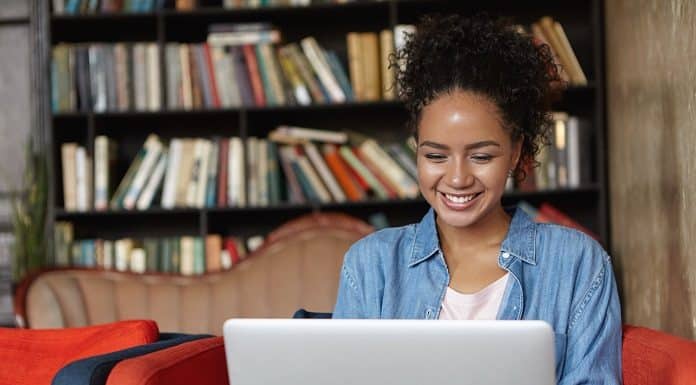 People, technology and education concept. Happy pretty dark skinned female in jean shirt sitting at sofa using laptop, typing something and browsing internet while being in reading hall or library.
